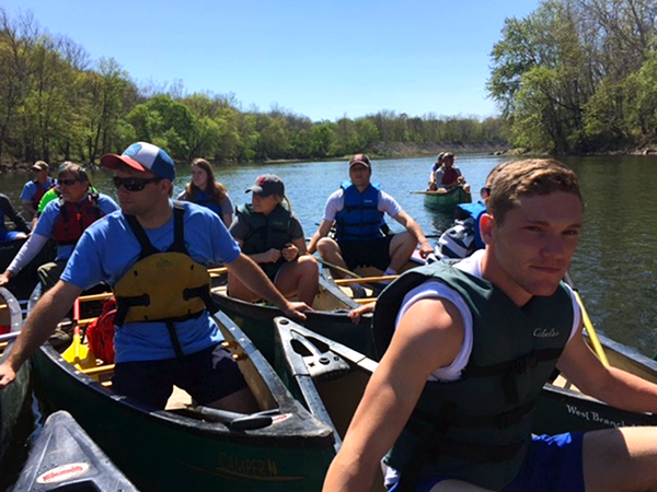 A cluster of canoes full of students wearing life vests float along a river with grassy, tree-filled banks and a blue sky.